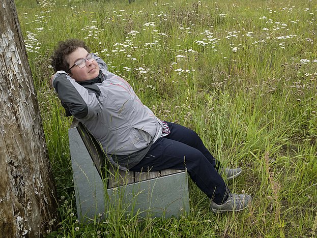 Eine Frau sitzt mitten in einer Blumenwiese auf einer Parkbank und blickt entspannt in den Himmel