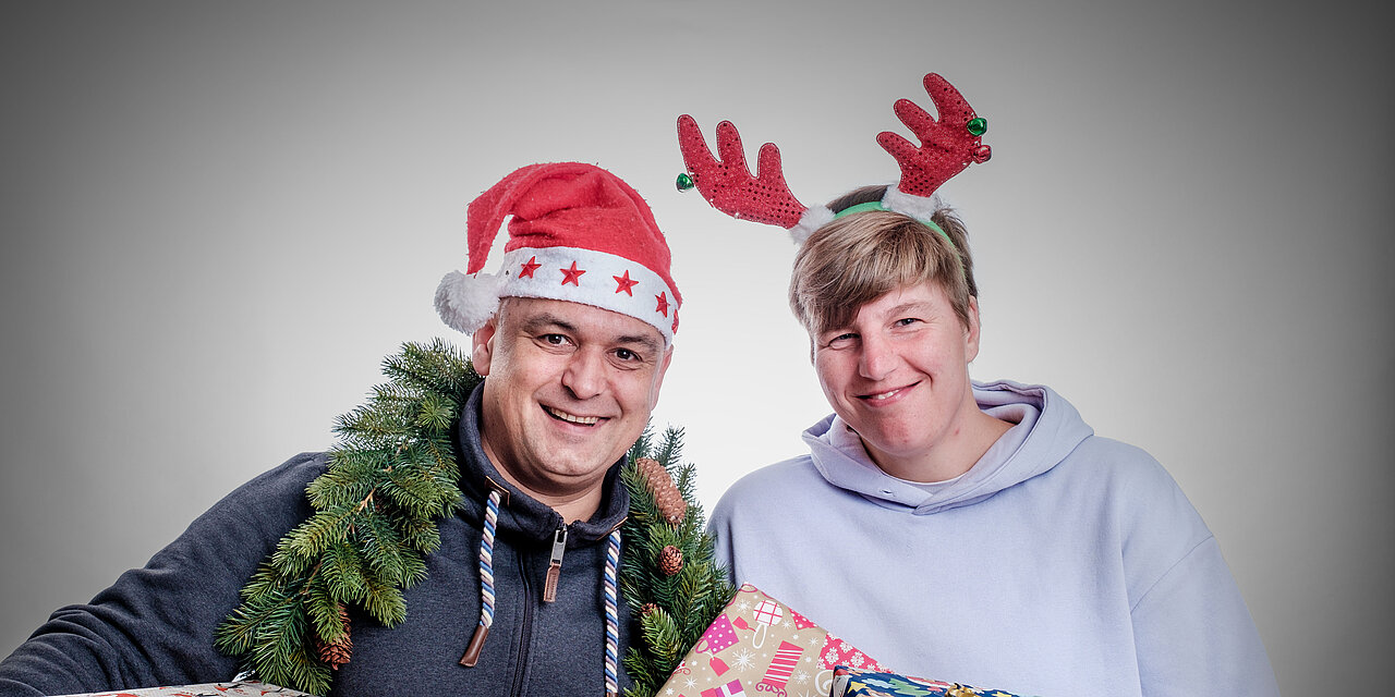 Zwei Menschen tragen Weihnachtsmütze und Elchgeweih auf ihren Köpfen, halten Geschenke in der Hand und lächeln in die Kamera 