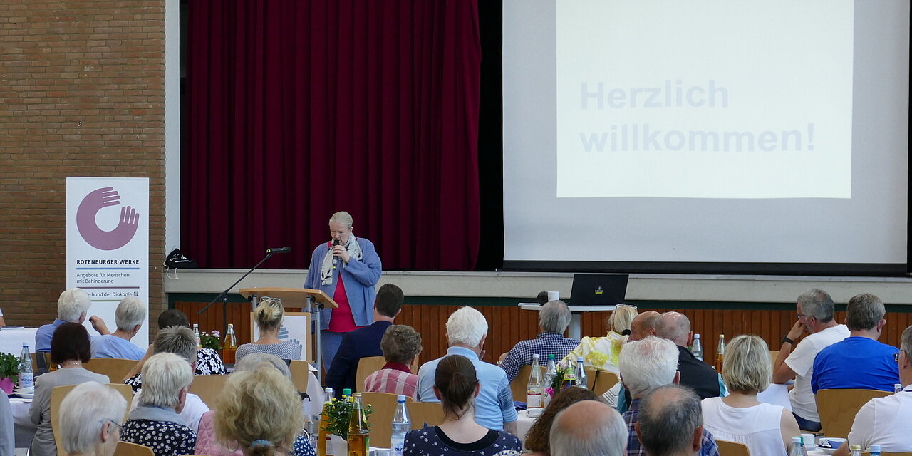 Menschen sitzen in einem Saal an Tischen und lauschen der Worten von Sabine Ulrich, die zu den Gästen spricht