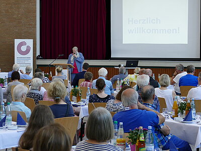 Menschen sitzen in einem Saal an Tischen und lauschen der Worten von Sabine Ulrich, die zu den Gästen spricht
