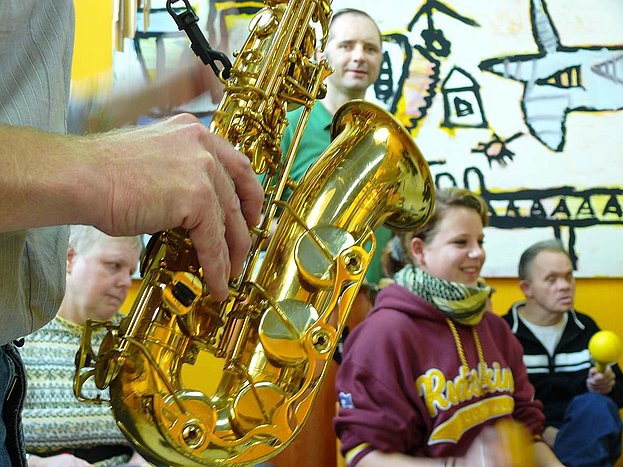 Ein Saxophon wird im Vordergrund gespielt, dahinter lächeln Menschen zur Musik