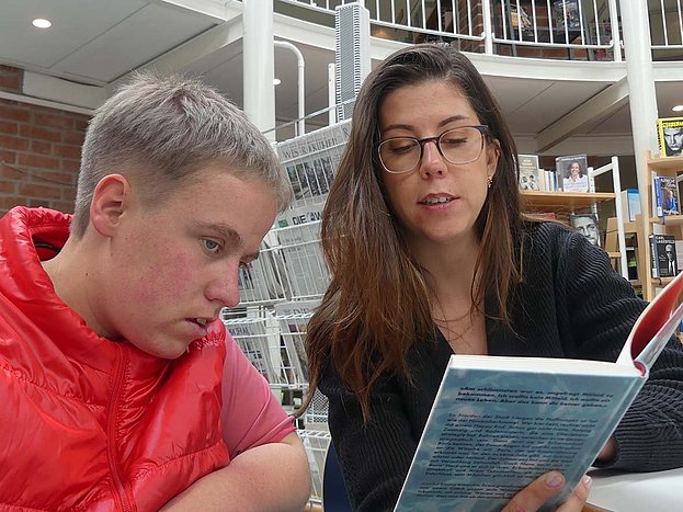Zwei junge Frauen - eine Bewohnerin und eine Mitarbeiterin sitzen in der Bücherei und betrachten ein Buch