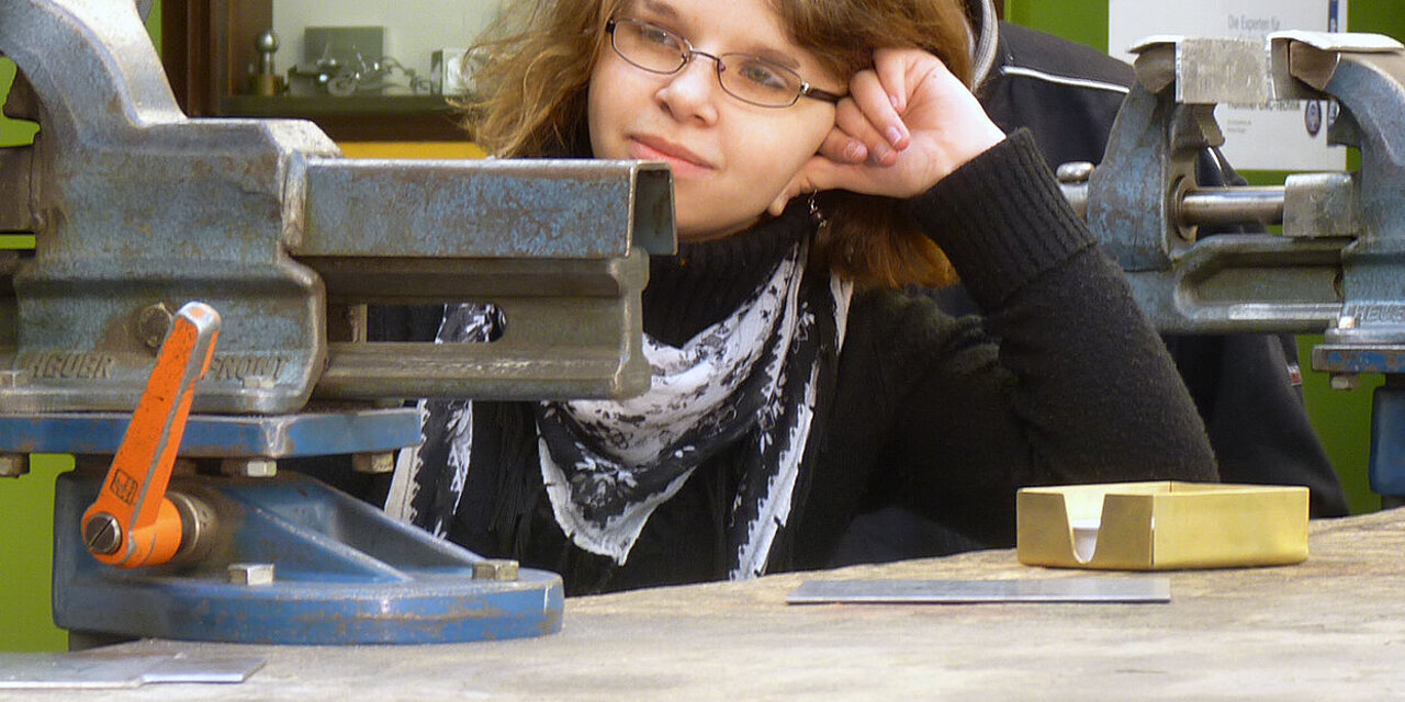 Eine junge Frau in einer Werkstatt stützt ihren Kopf an einer Werkbank ab