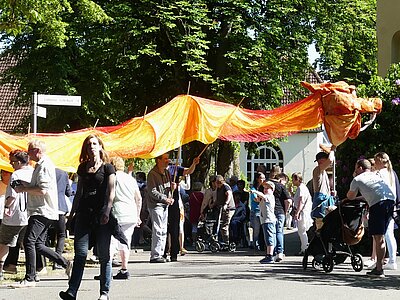 Sommerfest auf dem Kalandshof mit vielen Menschen bei sonnigem Wetter, eine große Drachenfigur aus Tüchern wird von mehreren Leuten getragen
