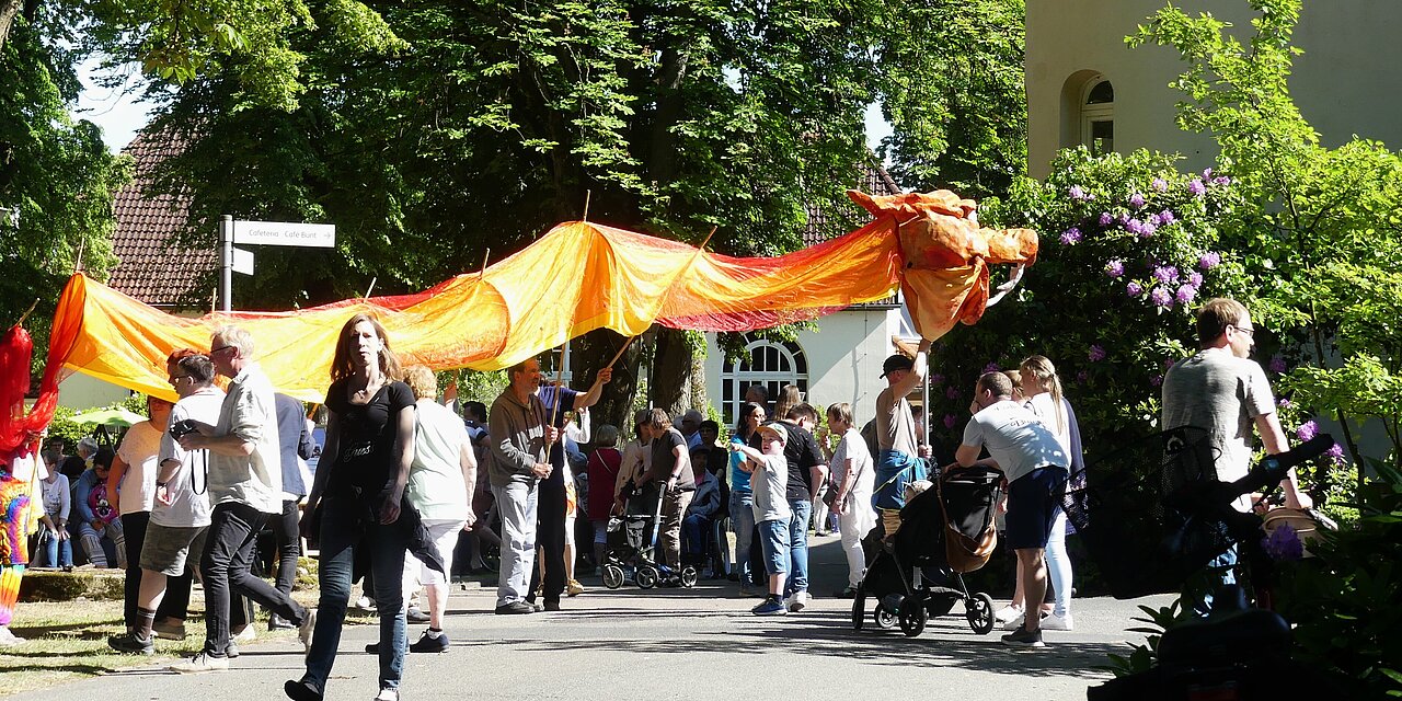 Sommerfest auf dem Kalandshof mit vielen Menschen bei sonnigem Wetter, eine große Drachenfigur aus Tüchern wird von mehreren Leuten getragen