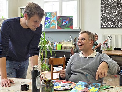In einem Kunstatelier sitzt ein Bewohner am Tisch und malt. Ein junger Mitarbeiter lehnt sich auf die Tischplatte und spricht mit ihm