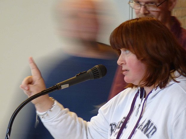 Drei Menschen stehen an einem Rednerpult, eine Frau spricht ins Mikrophon