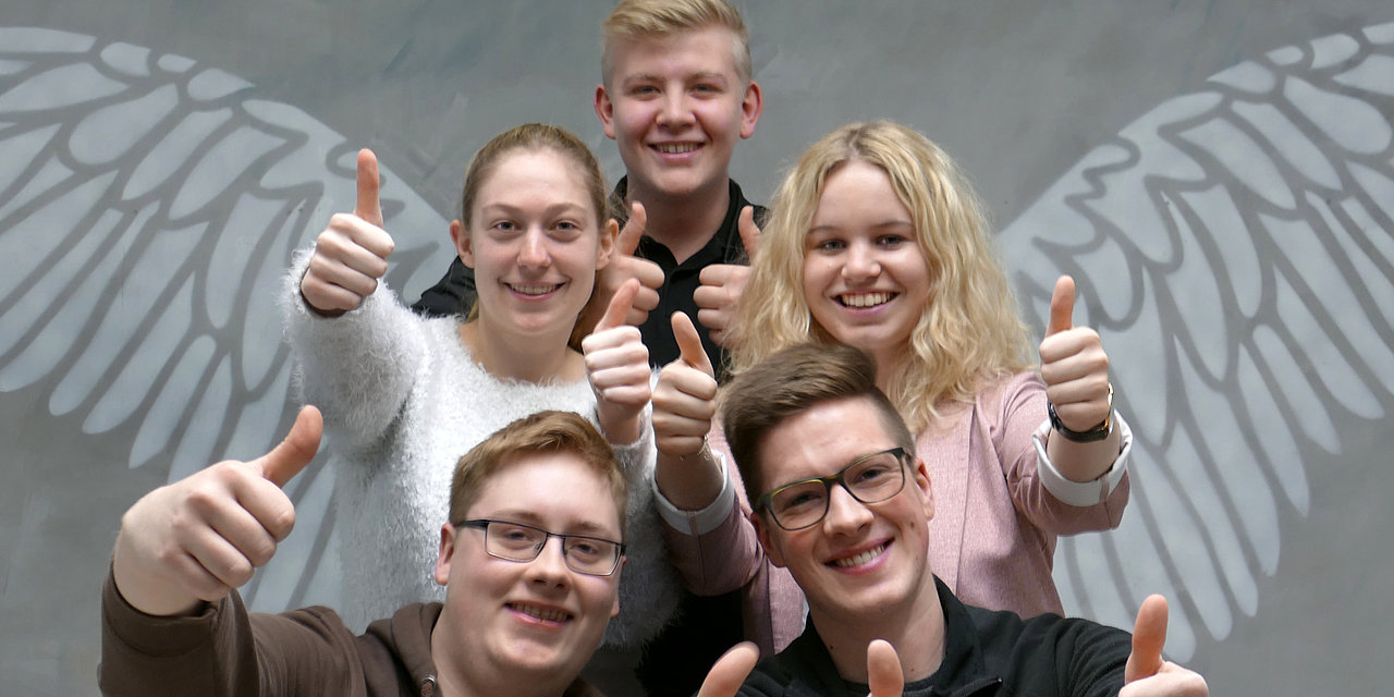 Fünf junge Leute stehen vor Engelsflügeln, haben ihre Daumen erhoben und lächeln in die Kamera