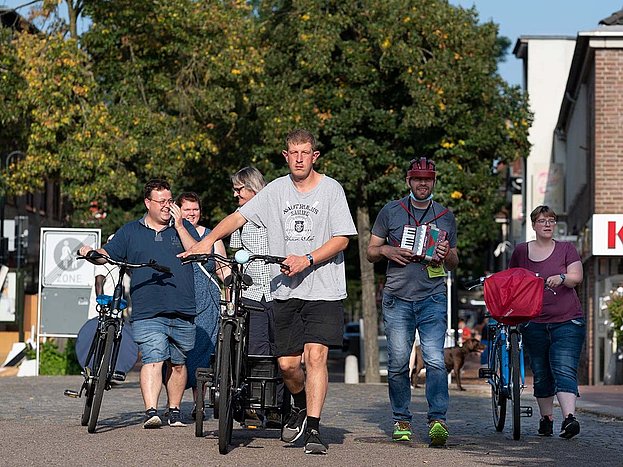 Mehrere Menschen schieben ihr Rad durch die sommerliche Fußgängerzone und gehen auf die Kamera zu