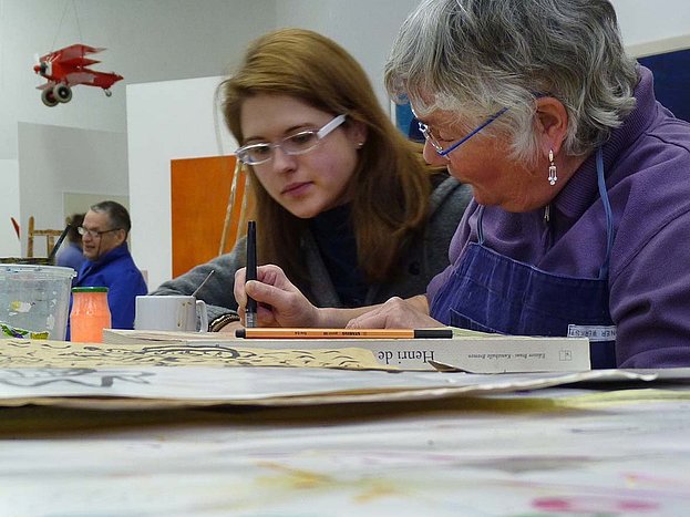 Zwei Frauen arbeiten konzentriert an einem Bild, das vor ihnen auf dem Tisch liegt
